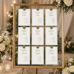 Peut inclure: Un plan de table en verre avec un cadre doré présentant neuf cartes blanches numérotées de Table One à Table Nine. Chaque carte affiche une liste de noms et est fixée en haut par un sceau en cire vert sauge orné d'un motif botanique.