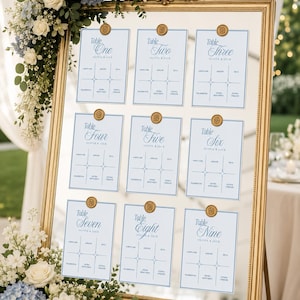 Puede incluir: Un espejo con marco dorado que muestra nueve tarjetas de asiento de boda blancas con texto azul, cada una con un sello de cera dorado en la parte superior. Las tarjetas están etiquetadas de Table One a Table Nine, con los nombres Olivia & Jack.