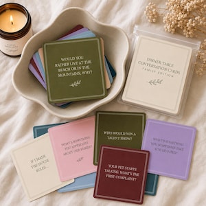 May include: A set of square conversation cards in various colors including olive green, pink, purple, and cream. The cards feature printed questions like 'Would you rather live at the beach or in the mountains, why?' and 'Who would win a talent show?'.