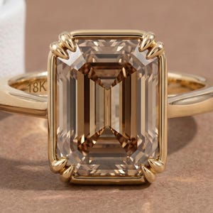 May include: A gold ring featuring a large, rectangular, emerald-cut champagne-colored gemstone set in a four-prong setting. The band is smooth and polished gold, with the engraving 18K visible on the inner surface of the ring shank.
