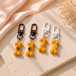 May include: Four keychains featuring yellow dog bone charms with paw print details. Two keychains have dark brown metal lobster clasps and rings, while the other two have white metal lobster clasps and rings. The charms are smooth and glossy.