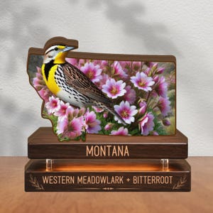 May include: A wooden display featuring a Western Meadowlark bird and pink Bitterroot flowers within a Montana-shaped cutout. The base is composed of two dark wood tiers with the text MONTANA and WESTERN MEADOWLARK + BITTERROOT engraved.
