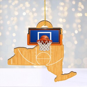 May include: A hanging ornament shaped like the state of New York, featuring a light wood grain finish with white basketball court lines. A rectangular blue backboard with an orange rim and a white net holds an orange basketball in the center.