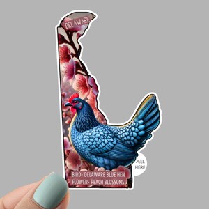 May include: A sticker in the shape of Delaware, featuring a blue hen with red comb and wattle, surrounded by pink peach blossoms. The text "DELAWARE" and "PEEL HERE" are visible. The text "BIRD- DELAWARE BLUE HEN FLOWER- PEACH BLOSSOMS" is also present.
