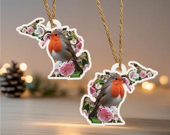 May include: Two wooden ornaments shaped like the state of Michigan, each featuring a robin perched on a floral branch. The robins have orange breasts and gray wings. The ornaments hang from a natural twine.