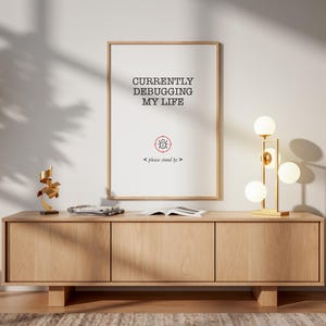 May include: A wooden framed print with the text "CURRENTLY DEBUGGING MY LIFE" is displayed above a wooden cabinet. A gold lamp with spherical bulbs and a small sculpture sit on the cabinet. The room has a modern, minimalist aesthetic.