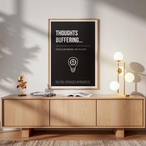 May include: A framed black and white poster with the text "THOUGHTS BUFFERING..." hangs above a light wood cabinet. A gold lamp with spherical bulbs and a modern sculpture sit on the cabinet. A wooden chair is to the right.
