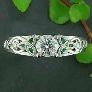 Pode incluir: Um anel de tom prateado com uma pedra central grande, redonda e transparente. A banda apresenta um trabalho de metal intrincado com um padrão de nós, acentuado com pequenas pedras redondas verdes incrustadas em ambos os lados da pedra.