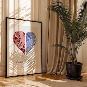 Puede incluir: Lámina artística enmarcada con un diseño en forma de corazón dividido en dos mitades, roja y azul, que representan mapas de ciudades. La impresión, en un marco negro, tiene los nombres "Kartil & Katila" en cursiva en la parte inferior.