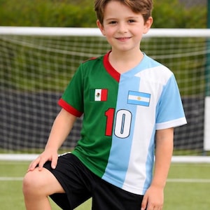 Pode incluir: Uma criança vestindo uma camisa de futebol dividida verticalmente com as bandeiras do México e da Argentina, com o número 10. Ele está em pé em uma bola de futebol, usando shorts pretos e tênis brancos.