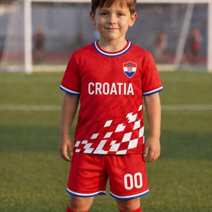 以下が含まれることがあります： 前面に「CROATIA」の文字がプリントされた赤いサッカーユニフォームを着た子供。ユニフォームは、赤色のジャージ、ショーツ、ソックスで構成され、白と青のアクセントが付いています。ショーツには番号「00」が表示されています。