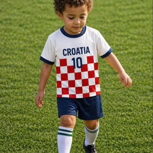 Puede incluir: Un ni&ntilde;o con un uniforme de f&uacute;tbol de Croacia, que incluye una camiseta blanca con "CROACIA 10", un dise&ntilde;o a cuadros rojos y blancos, pantalones cortos azul marino y calcetines blancos con rayas verdes. El ni&ntilde;o est&aacute; en un campo verde con un bal&oacute;n de f&uacute;tbol.