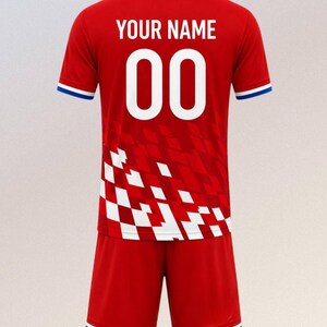 以下が含まれることがあります： 赤色のサッカーユニフォームとショーツのセット。ジャージには、白で「YOUR NAME」の文字と数字「00」が表示されています。ジャージとショーツには青と白のトリムが付いています。ジャージは赤と白のチェック柄です。