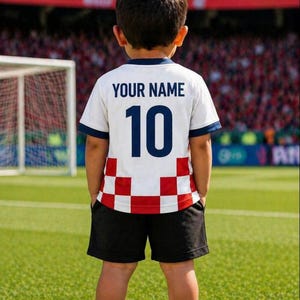 Puede incluir: Un ni&ntilde;o lleva una camiseta de f&uacute;tbol blanca con el texto "YOUR NAME" y el n&uacute;mero 10 en azul marino. La camiseta tiene un dise&ntilde;o a cuadros rojos y blancos en la parte inferior. El ni&ntilde;o lleva pantalones cortos negros y calcetines blancos.