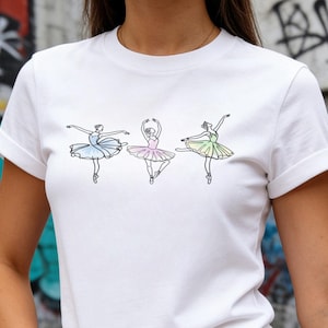 May include: White t-shirt featuring three ballet dancers in a line drawing style. Each dancer is wearing a tutu in a different pastel color: blue, pink, and green. The shirt has short sleeves and a crew neck.