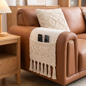 May include: A cream-coloured macrame sofa armrest organiser featuring a woven geometric pattern, a flat top section, and a hanging pocket with fringe tassels. Two black remote controls are tucked inside the pocket, resting on a brown leather sofa.