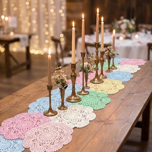 Könnte beinhalten: Eine Reihe runder, gehäkelter Spitzendeckchen in Pastelltönen wie Rosa, Blau, Gelb, Grün und Weiß, die in einer Linie auf einem Holztisch angeordnet sind. Das Arrangement umfasst mehrere hohe, schlanke Messingkerzenhalter mit brennenden Kerzen.
