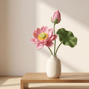 Op de afbeelding: Een gehaakt lotusbloemarrangement met een volledig bloeiende roze bloem met een geel hart, een gesloten roze knop en een enkel groen blad aan stelen. De bloemen staan in een gladde, matte, gebroken witte keramische vaas.