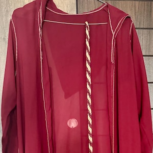 May include: A long, flowing burgundy robe with white stitching along the edges. A decorative cord with tassels hangs down the front. The robe is made of a lightweight, sheer fabric.