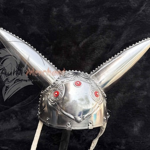 May include: A silver-colored helmet with two long, pointed ear-like projections. The helmet features intricate embossed designs and red circular accents. A white strap hangs from the bottom. The helmet is likely for costume or display.