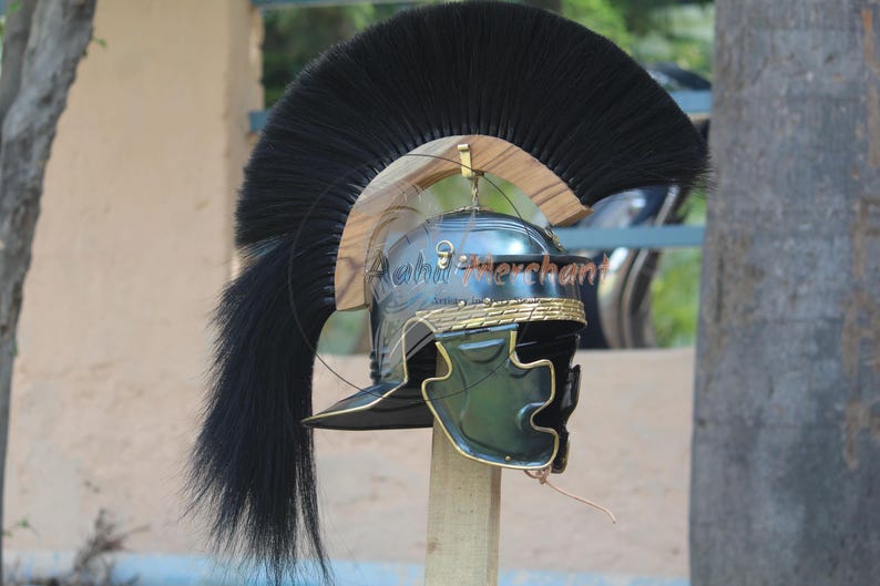 May include: A Roman helmet with a black plume. The helmet is dark green and gold with a curved visor and a decorative band around the base. The plume is long and flowing, adding to the helmet's imposing appearance.