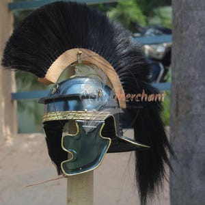 May include: A Roman helmet with a black plume. The helmet is primarily dark blue with gold trim and accents. The helmet is mounted on a wooden stand, showcasing its historical design and craftsmanship.