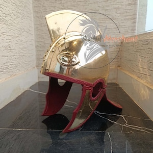 May include: A polished brass helmet with a red trim and accents. The helmet features a curved crest and is adorned with rivets and decorative elements. The design is reminiscent of ancient military headwear.