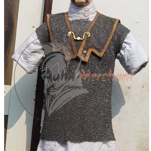 Puede incluir: Un chaleco de cota de malla con cuello de cuero marrón y cierre dorado. El chaleco se lleva sobre una camisa blanca con cuello alto. La imagen muestra una prenda medieval o histórica.