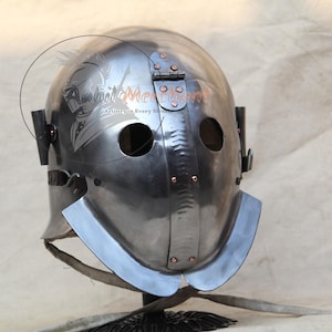 Puede incluir: Un casco de acero pulido con dos agujeros para los ojos y una parte superior con bisagras. El casco tiene una forma redondeada con un protector de cuello y una correa para la barbilla. El metal tiene una superficie reflectante y el casco parece ser de diseño histórico.