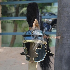 May include: A Roman helmet with a black plume and gold trim. The helmet is dark blue and black with a detailed design. The helmet is displayed outdoors, with a blurred background of trees and a window.