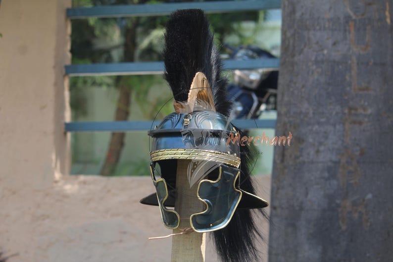May include: A Roman helmet with a black plume and gold trim. The helmet is dark blue and black with a detailed design. The helmet is displayed outdoors, with a blurred background of trees and a window.