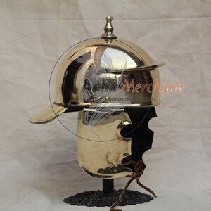 May include: A polished brass Roman helmet with a visor and a decorative finial. The helmet is mounted on a black stand with a detailed base. The image shows the helmet's reflective surface and intricate design.