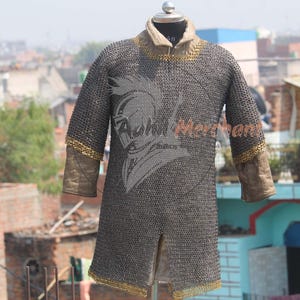 May include: A chainmail shirt displayed on a mannequin. The shirt is primarily gray with gold trim on the collar, cuffs, and hem. The sleeves are brown. A logo is visible on the chest.