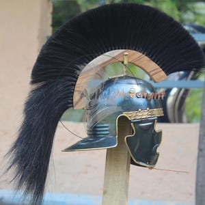 May include: A Roman-style helmet with a black plume and gold accents. The helmet is made of metal and features a face guard. The helmet is mounted on a wooden stand. The text "Achil Merchant" is visible on the helmet.