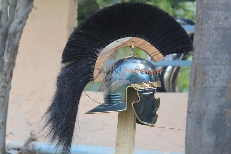 May include: A Roman-style helmet with a black plume and gold accents. The helmet is made of metal and features a face guard. The helmet is mounted on a wooden stand. The text "Achil Merchant" is visible on the helmet.