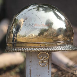 May include: A polished brass helmet with a rounded top and a decorative brim. The helmet has a textured surface and a gold-colored ring attached to the front. The helmet is displayed on a white post.