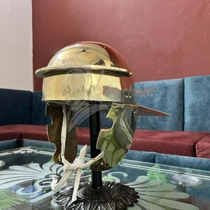 May include: A brass Roman helmet with a polished finish, displayed on a black stand. The helmet features a rounded top, a prominent brow guard, and cheek pieces. The background includes a red wall and a blue sofa.