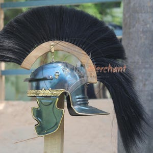 May include: A Roman helmet with a dark blue finish, featuring a prominent black plume. The helmet has gold-colored accents and a green cheek guard. It is mounted on a wooden stand, showcasing historical military headwear.