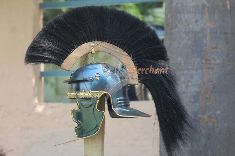 May include: A Roman helmet with a dark blue finish, featuring a prominent black plume. The helmet has gold-colored accents and a green cheek guard. It is mounted on a wooden stand, showcasing historical military headwear.