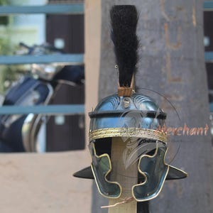 May include: A Roman helmet with a dark blue finish and gold-colored trim. The helmet features a black plume and is displayed on a wooden post. The helmet's design includes decorative elements and side cheek guards.