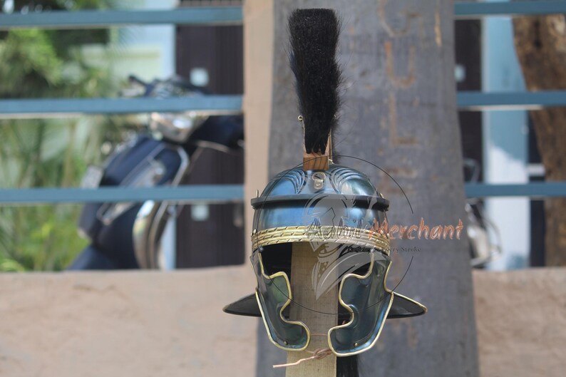 May include: A Roman helmet with a dark blue finish and gold-colored trim. The helmet features a black plume and is displayed on a wooden post. The helmet's design includes decorative elements and side cheek guards.