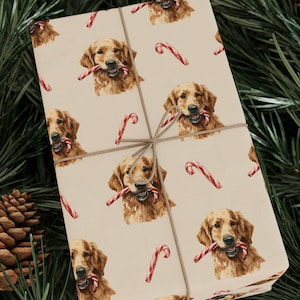 Puede incluir: Regalo envuelto con un fondo beige con un patrón repetido de golden retrievers sosteniendo bastones de caramelo. El regalo está atado con hilo y colocado sobre un fondo de agujas de pino y piñas.