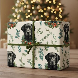 May include: A wrapped gift box featuring a repeating pattern of black Labrador Retrievers with festive greenery and red berries on a cream background. The box is tied with a green ribbon and bow, perfect for the holidays.