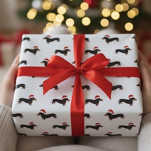 Puede incluir: Un regalo envuelto en papel blanco con un estampado de perros salchicha con gorros de Papá Noel. Está atado con una cinta y un lazo de satén rojo brillante. Un árbol de Navidad con luces borrosas está en el fondo.