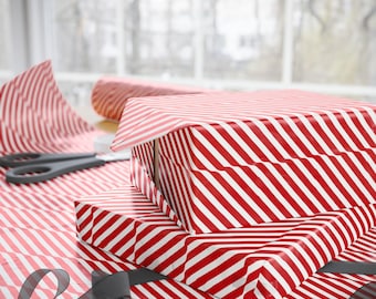 Puede incluir: Tres regalos envueltos en papel a rayas diagonales rojas y blancas están apilados sobre una mesa. También se ven un rollo de papel de regalo a juego, unas tijeras y una cinta gris, lo que sugiere una ocasión especial.