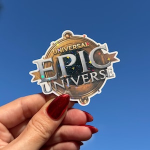 May include: A sticker with the words "UNIVERSAL EPIC UNIVERSE" in a circular design with gold and silver accents. The sticker is held up against a bright blue sky with palm trees in the background.