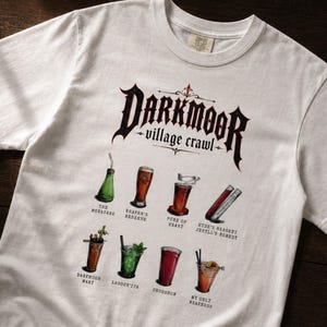 May include: A white short-sleeve t-shirt featuring a graphic print with the text Darkmoor Village Crawl at the top. Below the text are illustrations of eight different cocktails with labels including The Monocane, Reaper's Reserve, Pure of Heart, and Hyde's Reagent Jekyll's Remedy, Darkmoor Mary, Lagoon'ita, Crossbow, and My Only Weakness.
