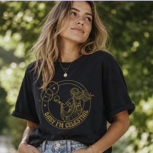 Puede incluir: Camiseta negra con mangas dobladas, con un gráfico dorado de una mujer sosteniendo una esfera, rodeada de estrellas y el texto "BABY I'M CELESTIAL". La camiseta es de un material suave.