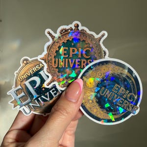 Puede incluir: Tres pegatinas holográficas con el texto "EPIC UNIVERSE". Las pegatinas tienen un diseño circular con un tema celestial, con engranajes, estrellas y una rosa de los vientos. Las pegatinas están sujetas por una mano.