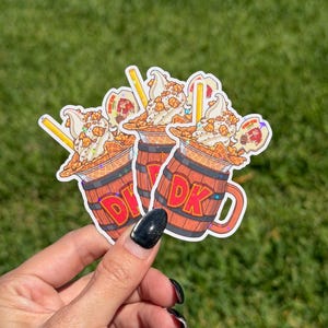 Puede incluir: Tres pegatinas con un helado de dibujos animados en una taza con forma de barril de madera. El helado tiene un remolino de helado suave, salsa de caramelo y una galleta de oblea. La taza tiene las letras "DK" en rojo.
